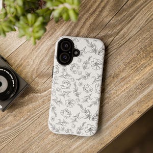 Puede incluir: Una funda de teléfono blanca con un estampado floral negro de flores y hojas. La funda tiene un recorte para una cámara de doble lente y está sobre una superficie de madera. Una cámara y vegetación son visibles en el fondo.
