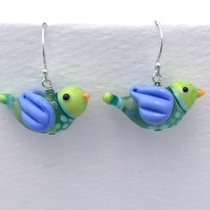 Puede incluir: Un par de pendientes de pájaro de cristal hechos a mano. Cada pendiente presenta un cuerpo verde con alas azules, una cabeza verde claro y un pico naranja. El cuerpo del pájaro está adornado con lunares blancos. Los pendientes cuelgan de ganchos plateados.