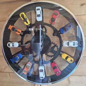 May include: A circular wall clock featuring a black gear-shaped frame with twelve miniature toy cars attached as hour markers. The clock has a silver rim, a clear glass face, and simple silver hands. The cars are in various colours including yellow, white, red, blue, and orange.