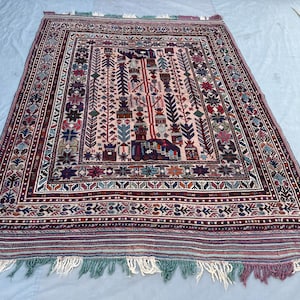 May include: A rectangular, hand-woven rug with a central design featuring geometric and floral patterns in shades of red, blue, green, and cream. The rug has a detailed border and fringed edges, showcasing traditional craftsmanship.