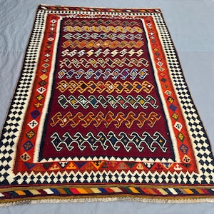 May include: A rectangular, hand-woven rug with a deep red central field. The rug features geometric patterns in various colors, including yellow, blue, green, and orange. The border has a black and white checkered pattern, with additional colorful geometric designs.