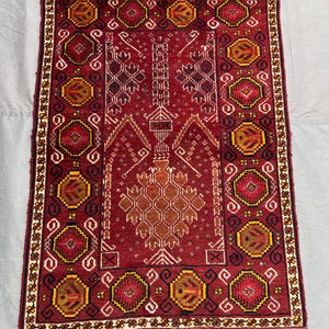 Può includere: Un tappeto rettangolare, tessuto a mano, con sfondo rosso intenso e intricati motivi geometrici. Il design presenta forme ottagonali ripetute in giallo, marrone e rosso, incorniciate da un bordo bianco e marrone. Il design centrale del tappeto include un motivo floreale stilizzato.