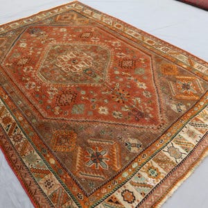 May include: A vintage rug with a red, orange, and brown color scheme. The rug has a central medallion with intricate floral and geometric patterns. The border is a cream color with a repeating pattern of stylized leaves and flowers.