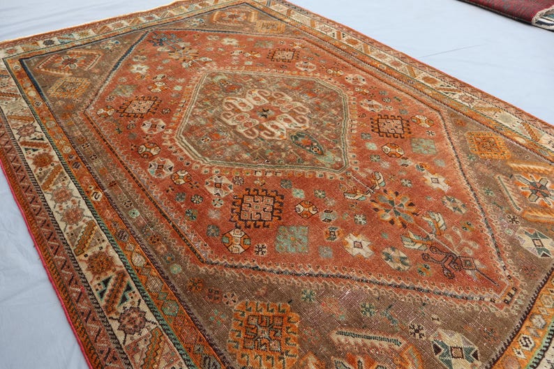 May include: A vintage rug with a geometric pattern in shades of red, orange, brown, and white. The rug has a central diamond shape with intricate details and a border with repeating motifs.