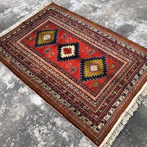 Pode incluir: Um tapete vermelho, verde e preto com franja branca. O tapete tem um design tradicional com formas geométricas.