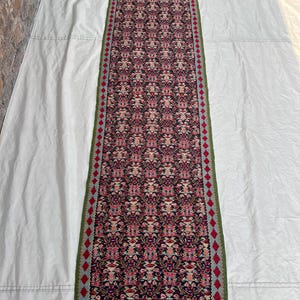 May include: A long, narrow rug with a repeating floral pattern in shades of red, pink, and cream on a dark background. The rug has a green border and a red diamond pattern along the edges. The rug is likely made of woven material.