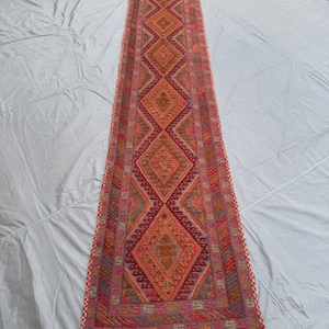 May include: A long, narrow runner rug with a repeating diamond pattern in shades of red, orange, and brown. The rug has a detailed geometric design and a fringed edge. The runner is likely made of woven material.