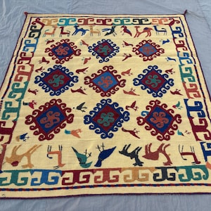 May include: A rectangular, cream-colored rug with a repeating pattern of stylized animals, birds, and geometric shapes in red, blue, green, and brown. The rug has a border with a repeating design and tassels on the corners.