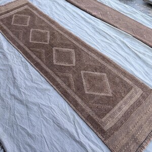 May include: A rectangular brown rug with a diamond pattern. The rug has a fringe on the short ends and a border. The diamond pattern is repeated four times down the center of the rug. The rug is made of a woven material.