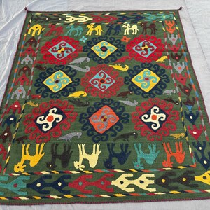 Könnte beinhalten: Ein rechteckiger, handgewebter Teppich mit dunkelgrünem Hintergrund. Der Teppich weist ein sich wiederholendes Muster aus geometrischen Formen, Tieren und floralen Motiven in Rot, Gelb, Blau und Weiß auf. Der Rand hat ein sich wiederholendes Muster aus stilisierten Tieren und Formen.
