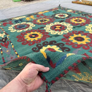 Könnte beinhalten: Handgewebter, quadratischer Teppich mit türkisgrünem Hintergrund und einem sich wiederholenden Blumenmuster in Gelb, Rot und Weiß. Der Teppich hat einen detaillierten Rand und Quasten an den Ecken. Der Teppich besteht aus dickem, strapazierfähigem Material.