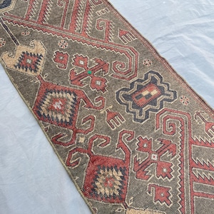 May include: A rectangular, patterned rug with a muted olive-green background. The rug features intricate geometric designs in shades of red, tan, and black. The design includes repeating shapes and patterns, creating a traditional, textured look.