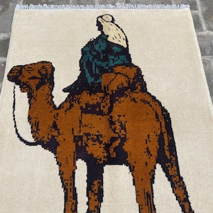 以下が含まれることがあります： ベージュの敷物に、茶色のラクダと緑と黒の服を着た人が乗っています。ラクダは画像の左側を向いています。