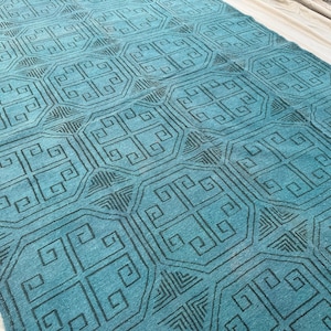 Puede incluir: Una alfombra turquesa con un patrón geométrico repetitivo en negro. El diseño presenta cuadrados entrelazados y formas estilizadas, creando una apariencia texturizada. La alfombra es rectangular y parece estar hecha de material tejido.