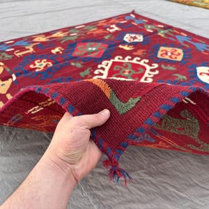 May include: A hand holds a section of a red, patterned rug. The rug features a repeating design with geometric shapes and figures in blue, yellow, and white. The edges are trimmed with blue and red stitching, and the rug appears to be made of woven fabric.