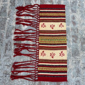 May include: A red, white, and green woven tapestry with fringe on the sides. The tapestry features a geometric pattern with stylized birds.