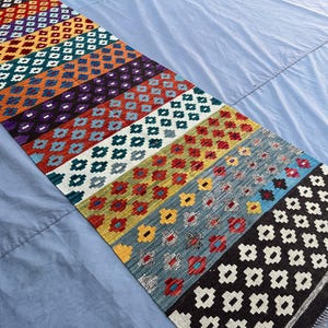 May include: A colorful runner rug with a geometric pattern. The rug features rows of diamonds and floral motifs in various colors, including red, blue, orange, and purple. The rug has a fringed edge.