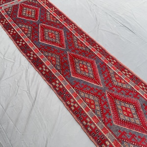 May include: A rectangular runner rug with a repeating diamond pattern in red and gray. The rug has a detailed border with small geometric shapes. The design is symmetrical and the rug appears to be made of woven material.