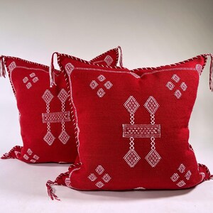 Puede incluir: Dos cojines decorativos rojos con patrones geométricos bordados en blanco. Los cojines tienen costuras blancas en los bordes y borlas en las esquinas. Los cojines son de forma cuadrada.