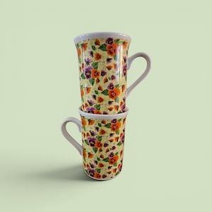 May include: Two stacked ceramic mugs with a floral pattern. The mugs have a yellow background with purple and orange flowers and green leaves. The rims and handles are white.