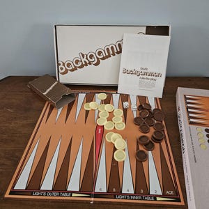 Puede incluir: Un juego de backgammon vintage con un tablero marrón y blanco, fichas beige y marrón, dados y un libro de reglas. La caja blanca tiene la inscripción "Backgammon". El juego está dispuesto sobre una superficie de madera.