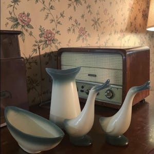 Puede incluir: Colección de decoración de cerámica vintage, que incluye dos figuras de gansos, un jarrón y un cuenco, todos con una combinación de colores blanco y verde azulado. Los artículos están colocados sobre una mesa de madera oscura, con una radio vintage al fondo.