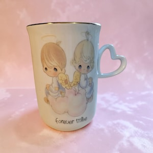 May include: Cream-colored ceramic mug with a gold rim and a heart-shaped handle. The mug features a charming illustration of two cherubic figures with halos and wings, seated on a cloud. The words "forever mine" are printed at the bottom.