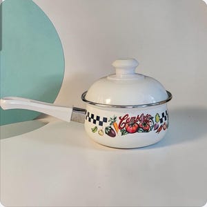 May include: A white enamel saucepan with a lid and a long handle. The pot features a black and white checkered border, a colorful vegetable graphic, and the red Campbell's logo printed on the side. The lid has a matching white knob handle.