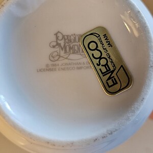 May include: Close-up of a white ceramic Precious Moments collectible. The bottom of the piece is visible, with the text "PRECIOUS MOMENTS" and "&copy; 1984 JONATHAN & D LICENSEE ENESCO IMPORT". A gold and black "ENESCO" sticker is also present.