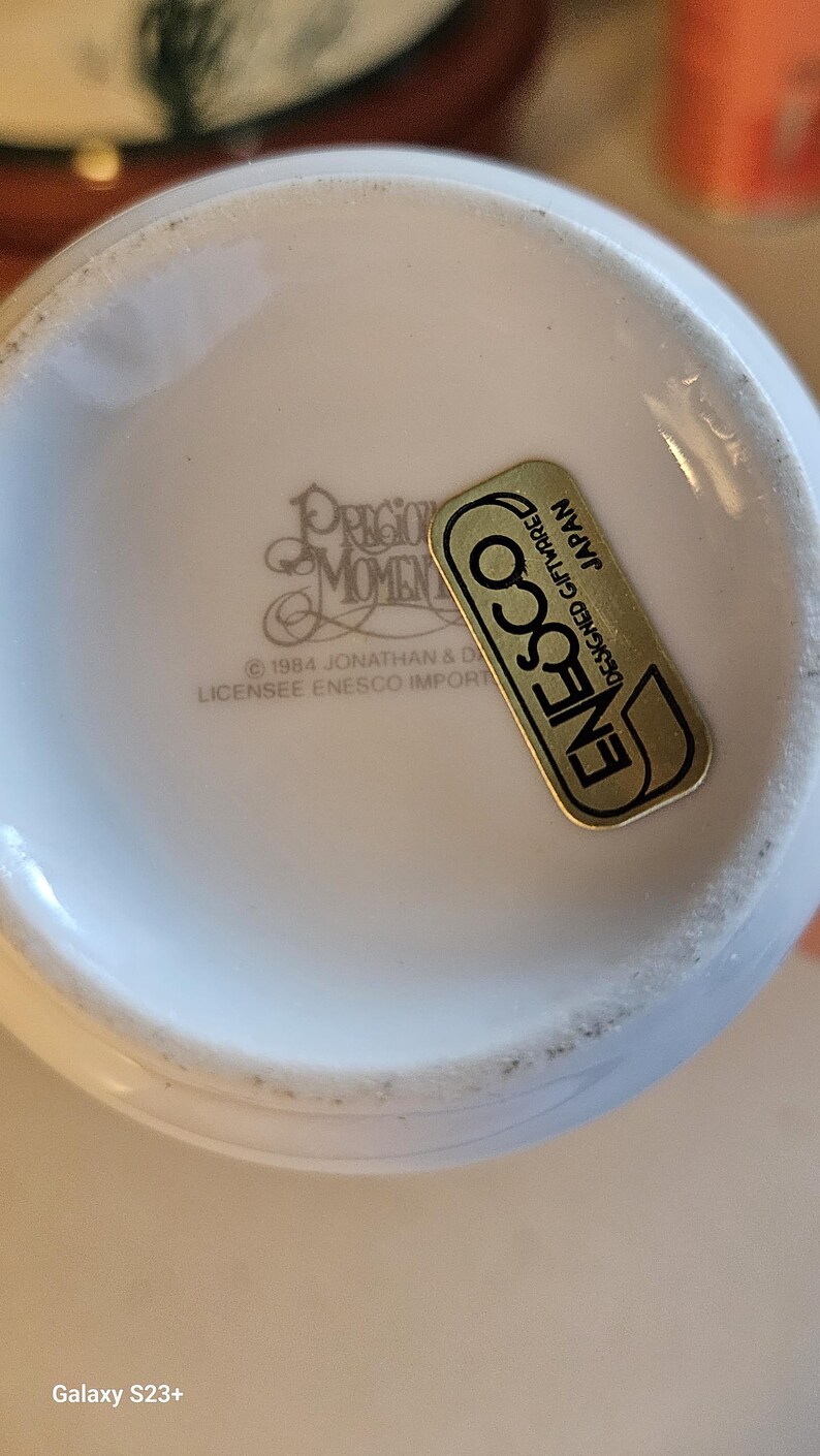 May include: Close-up of a white ceramic Precious Moments collectible. The bottom of the piece is visible, with the text "PRECIOUS MOMENTS" and "&copy; 1984 JONATHAN & D LICENSEE ENESCO IMPORT". A gold and black "ENESCO" sticker is also present.