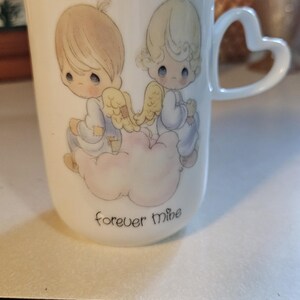 May include: White ceramic mug with a heart-shaped handle and gold trim. The mug features a design of two cherubic figures with wings, sitting on a cloud, with the words "forever mine" below. A collectible item.