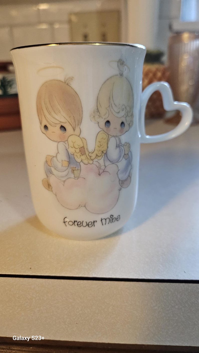 May include: White ceramic mug with a heart-shaped handle and gold trim. The mug features a design of two cherubic figures with wings, sitting on a cloud, with the words "forever mine" below. A collectible item.