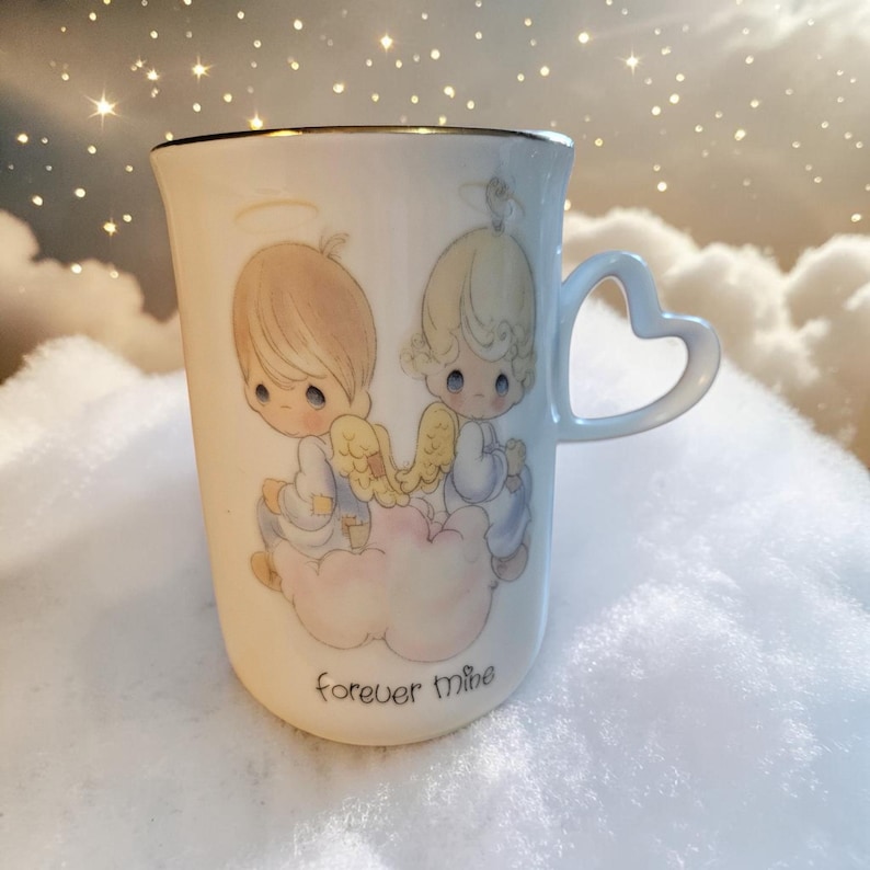 May include: Cream-colored ceramic mug with a gold rim and heart-shaped handle. The mug features a design of two cherubic figures with wings, sitting on a cloud. The words "forever mine" are printed on the front.