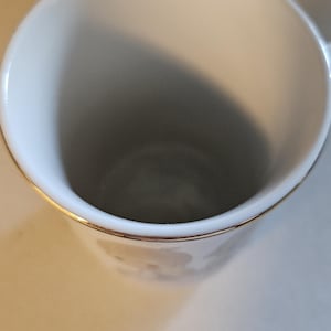 May include: A white ceramic mug with a gold rim. The interior of the mug is white, and the exterior features a subtle floral design. The mug is empty and sits on a light-colored surface.