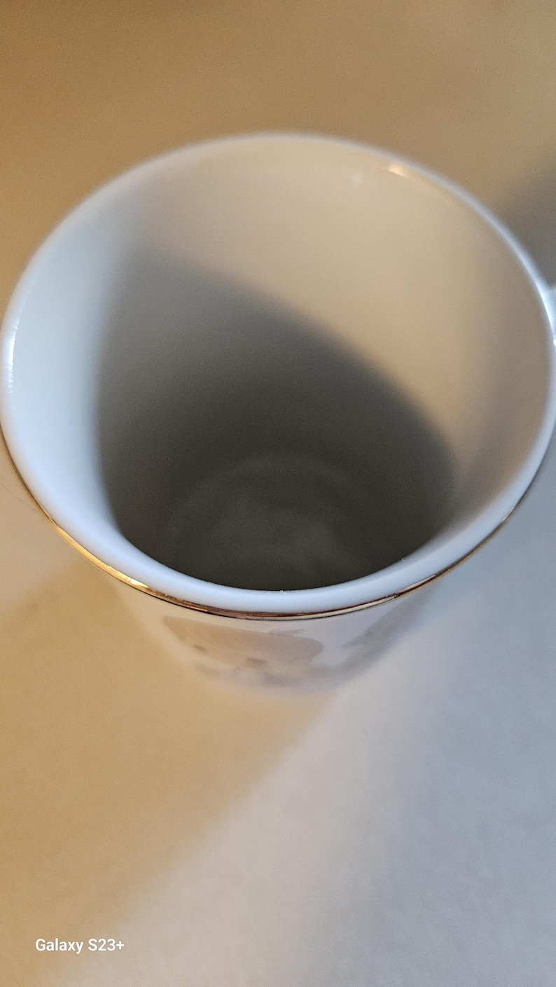 May include: A white ceramic mug with a gold rim. The interior of the mug is white, and the exterior features a subtle floral design. The mug is empty and sits on a light-colored surface.