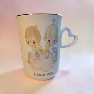 May include: A white ceramic mug with a gold rim and a heart-shaped handle. The mug features a design of two cherubic figures with halos and wings, sitting on a cloud. The words "forever mine" are printed on the mug.