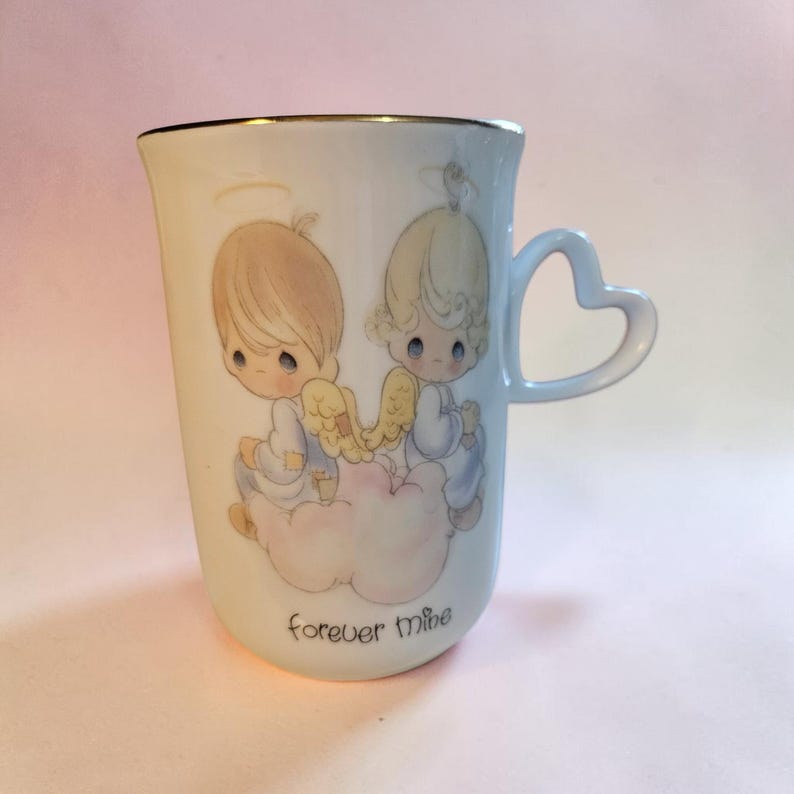 May include: A white ceramic mug with a gold rim and a heart-shaped handle. The mug features a design of two cherubic figures with halos and wings, sitting on a cloud. The words "forever mine" are printed on the mug.