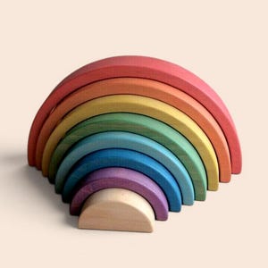 Puede incluir: Un juguete apilable de arcoíris de madera en tonos rojo, naranja, amarillo, verde, azul y morado. El juguete está hecho de varias piezas arqueadas que se encajan. La base es de color madera natural.