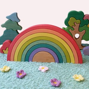 Pode incluir: Um brinquedo empilhável de arco-íris de madeira com arcos em várias cores, incluindo vermelho, laranja, amarelo, verde, roxo e madeira natural. O brinquedo é acompanhado por árvores e flores de madeira, tudo sobre uma superfície azul.