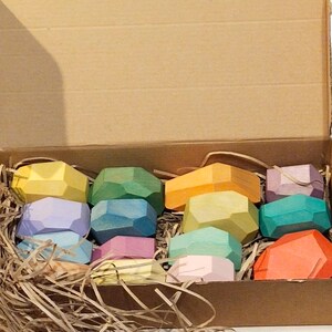 Könnte beinhalten: Eine Schachtel gefüllt mit bunten geometrischen Holzformen, die an Edelsteine erinnern. Die Formen sind in verschiedenen Schattierungen von Grün, Blau, Gelb, Orange, Rosa und Rot. Die Schachtel ist mit zerkleinertem Papier gefüllt.