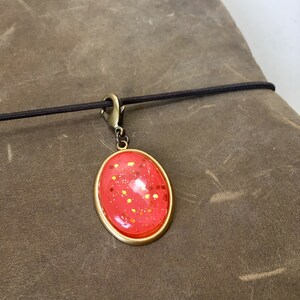 May include: Oval pendant with a red glitter design set in a gold-tone frame. The pendant is attached to a black elastic cord with a gold-tone clasp. The background is a textured brown surface.