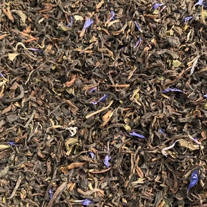 May include: Close-up of loose leaf tea with a mix of dark brown and green leaves, and small blue flower petals. The tea leaves are tightly packed together, creating a textured appearance. This image showcases a blend of tea, perfect for brewing.