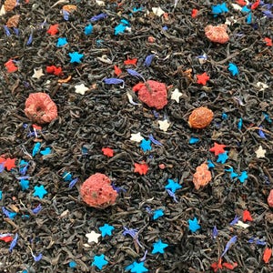 May include: Close-up of loose leaf tea with red, white, and blue star-shaped sprinkles, dried berries, and blue flower petals. The tea leaves are dark brown. A festive blend for a patriotic theme.