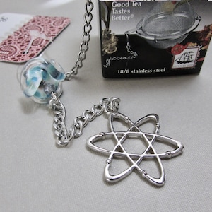 May include: A stainless steel tea infuser with a chain and a decorative atom-shaped charm. The infuser includes a glass bead with blue and white swirls. The packaging reads "Good Tea Tastes Better".