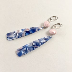 May include: A pair of dangle earrings. Each earring features a long, marbled blue, white, and pink teardrop shape, connected to a light pink faceted bead and a silver-colored lever-back clasp. The earrings are set against a white background.