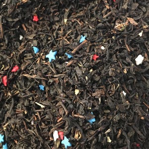 May include: Close-up of loose leaf tea with colorful star-shaped sprinkles. The tea leaves are dark brown, and the sprinkles are red, white, and blue. The tea is likely a flavored blend, perfect for a patriotic theme.