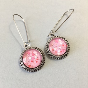 May include: A pair of dangle earrings with a pink glitter design. The earrings have a round, silver-colored frame with a textured edge. The ear wires are silver-colored and curved.