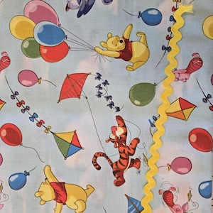 Op de afbeelding: Een blauwe stof met een herhalend patroon van Winnie de Poeh-karakters, vliegers en ballonnen. De karakters zijn Winnie de Poeh, Tigger, Biggetje en Eeyore. De stof is perfect voor quilten, knutselen en andere naaiprojecten.
