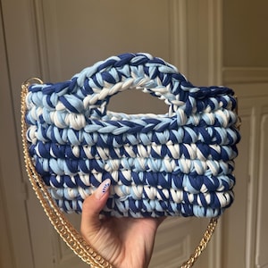 Könnte beinhalten: Handgefertigte, blau-weiß gehäkelte Handtasche mit goldener Kette. Die Tasche hat eine rechteckige Form mit einem stabilen Griff und einer Mischung aus dunkelblauem, hellblauem und weißem Garn. Die goldene Kette sorgt für einen eleganten Touch.