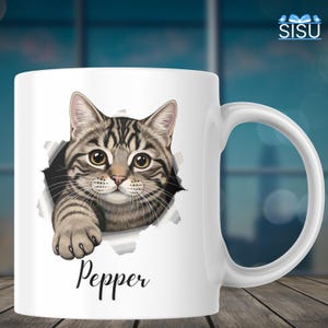 May include: White ceramic mug featuring a tabby cat design. The cat's face and paw appear to be emerging from a torn hole. The name "Pepper" is written in cursive below the cat. The mug has a curved handle.